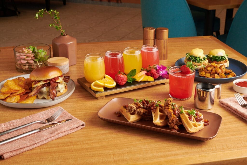Brunch spread at Honey + Vine featuring mimosas, pancakes, burgers and breakfast dishes on a wooden table in a modern dining setting.