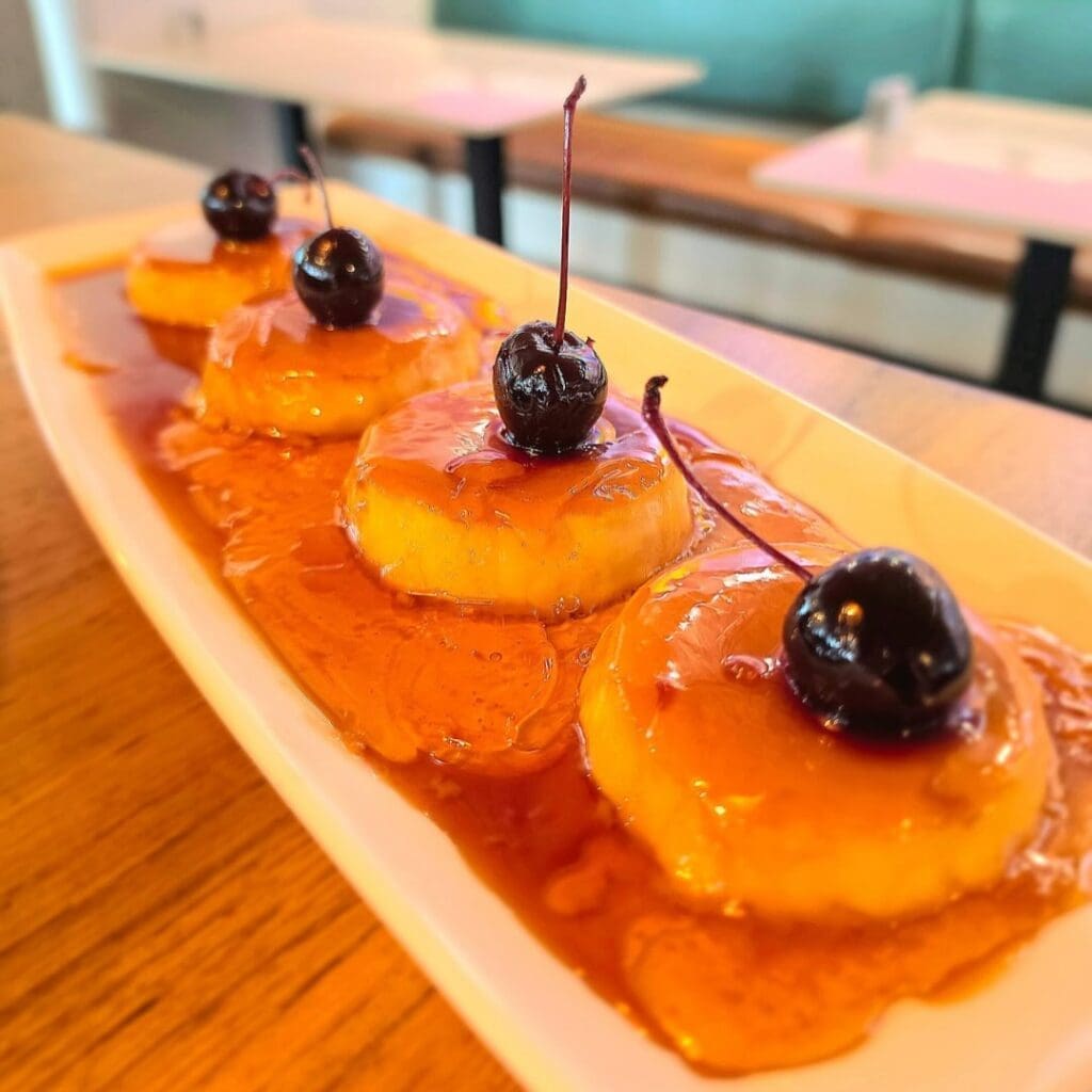 Close-up of caramel flan topped with cherries and caramel sauce on a rectangular plate at a restaurant table.