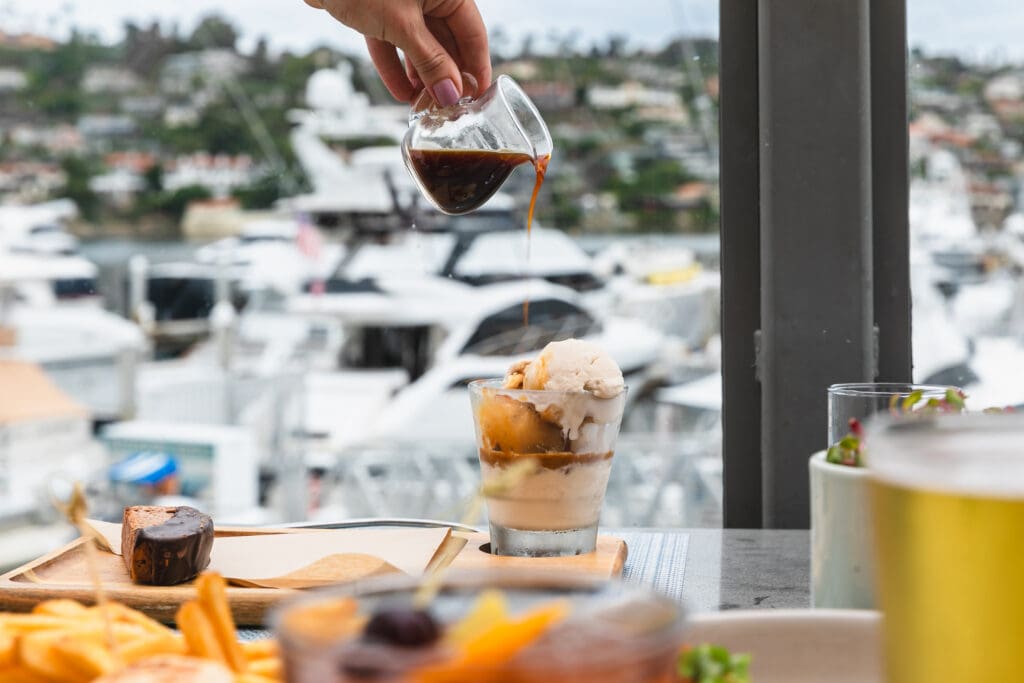 A hand poors coffee into an ice cream making an afagado with harbor boats in the background at Vessel Restaurant. 