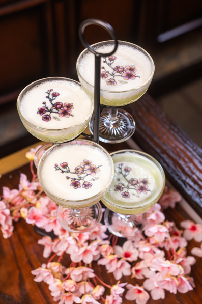 Tiered tower of elegant cocktails garnished with delicate floral details, surrounded by pink blossoms on a wooden table.