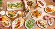 Overhead view of a colorful brunch spread with flatbreads, tacos, sliders, fries and cocktails on a shared table.