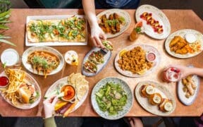 Overhead view of a colorful brunch spread with flatbreads, tacos, sliders, fries and cocktails on a shared table.