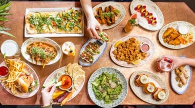 Overhead view of a colorful brunch spread with flatbreads, tacos, sliders, fries and cocktails on a shared table.