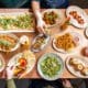 Overhead view of a colorful brunch spread with flatbreads, tacos, sliders, fries and cocktails on a shared table.