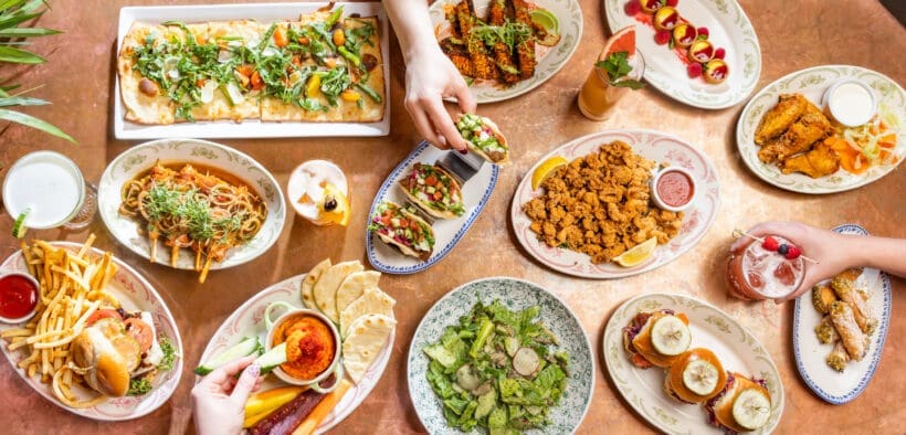 Overhead view of a colorful brunch spread with flatbreads, tacos, sliders, fries and cocktails on a shared table.