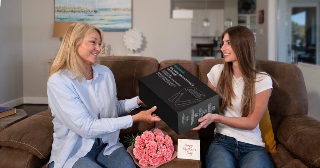 A woman gifting a neck and back massager to another woman sitting on a couch with flowers and a Mother’s Day card nearby.