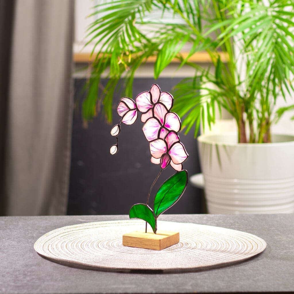 A stained glass pink orchid tabletop sculpture on a woven placemat with a green plant in the background.