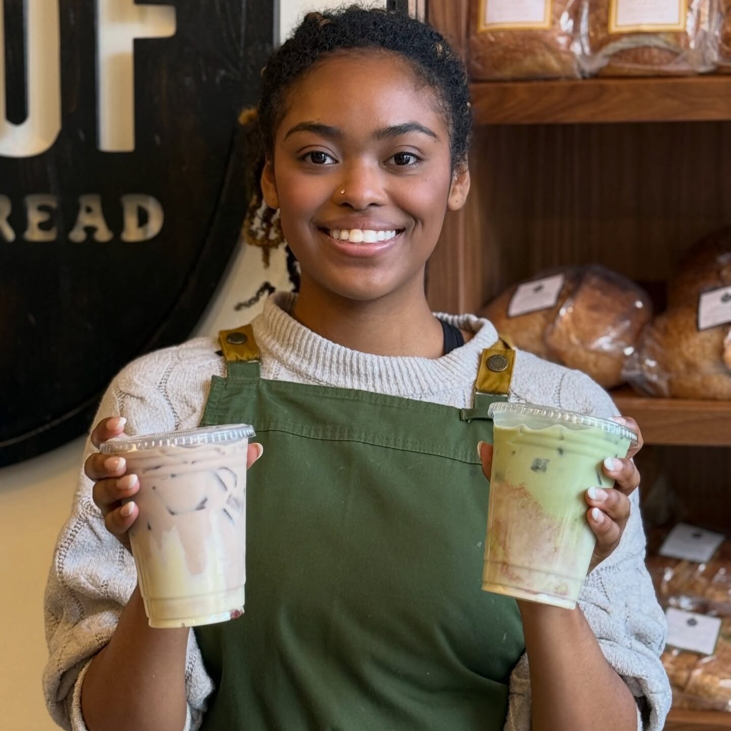 Barista holding two iced drinks inside a bakery with shelves of fresh bread behind them.