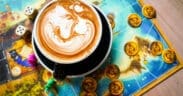 Latte with detailed foam art sitting on a colorful board game with dice and game pieces at nakes & Lattes.