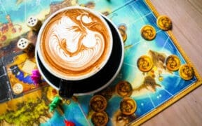 Latte with detailed foam art sitting on a colorful board game with dice and game pieces at nakes & Lattes.