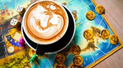 Latte with detailed foam art sitting on a colorful board game with dice and game pieces at nakes & Lattes.