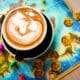 Latte with detailed foam art sitting on a colorful board game with dice and game pieces at nakes & Lattes.
