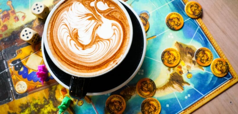 Latte with detailed foam art sitting on a colorful board game with dice and game pieces at nakes & Lattes.