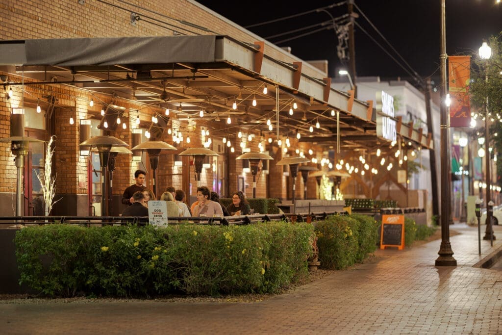 Outdoor patio at night with string lights, people seated at tables and a brick building exterior at Snakes and Lattes in Downtown Tempe.