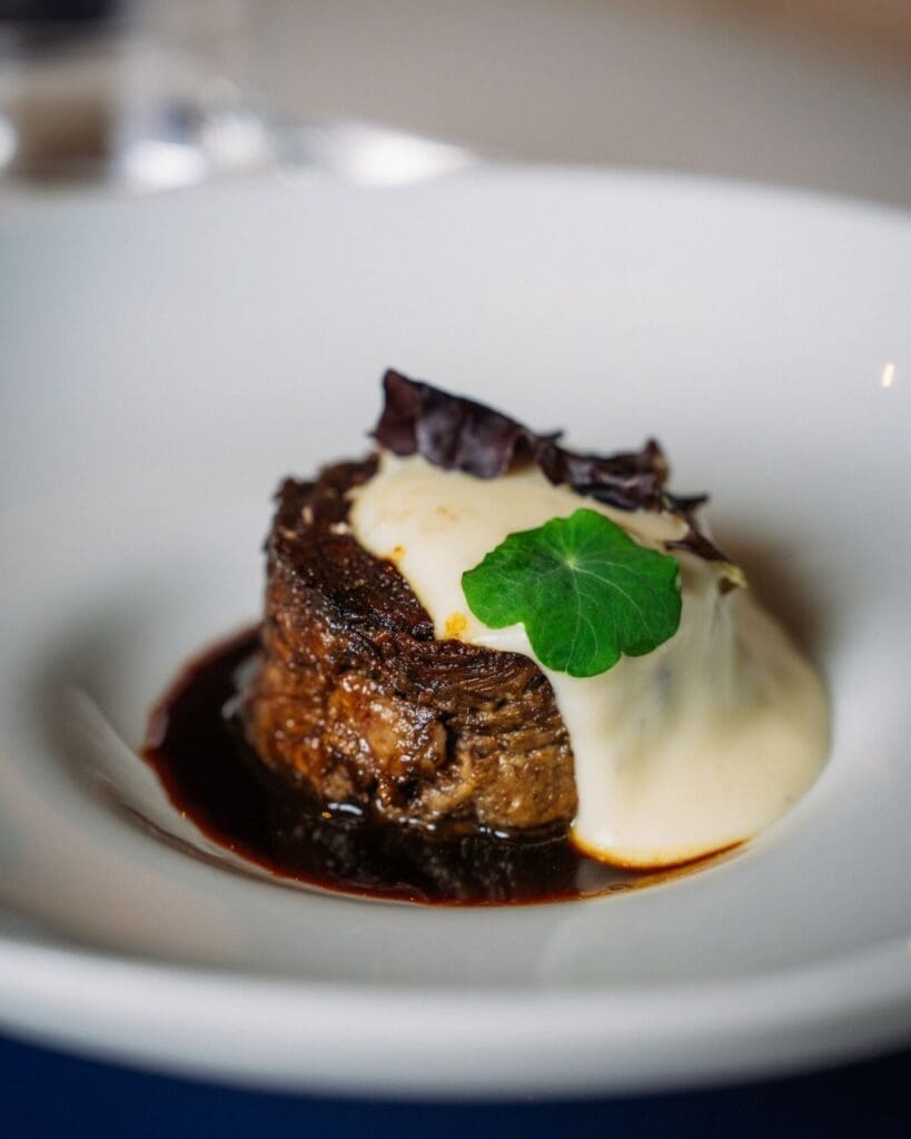 Close-up of a plated braised beef entrée topped with creamy sauce and greens in a refined restaurant setting.