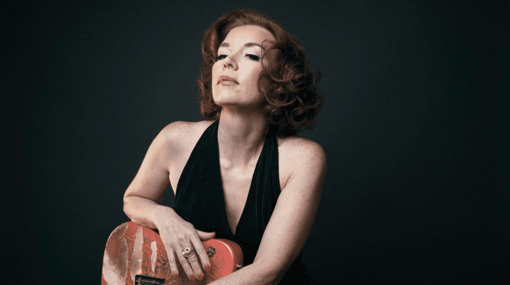 Portrait of blues guitarist Sue Foley holding guitar against dark background ahead of live performance.
