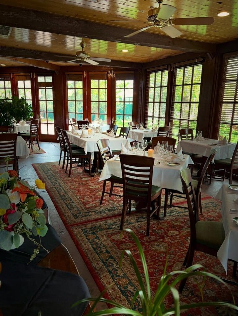 Elegant dining room at The Greenbriar Inn with white tablecloth tables, wood-paneled ceilings, large windows and natural light overlooking greenery.