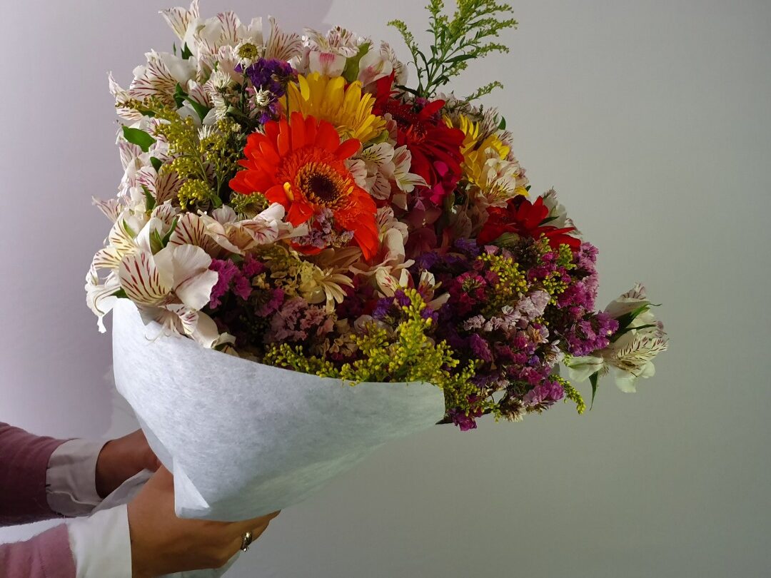 Hand holding a vibrant spring bouquet with orange, yellow and pink flowers against a neutral background.