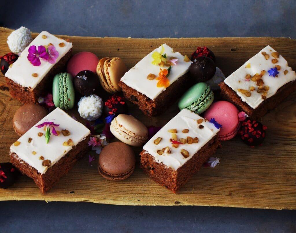 Assorted desserts including frosted cake squares, colorful macarons and chocolate truffles arranged on a rustic wooden board.