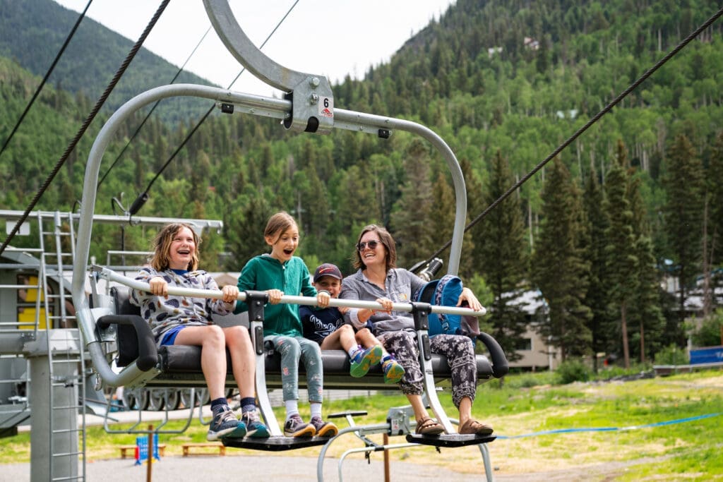 Family riding a scenic chairlift surrounded by forested mountain views at Taos Ski Valley.