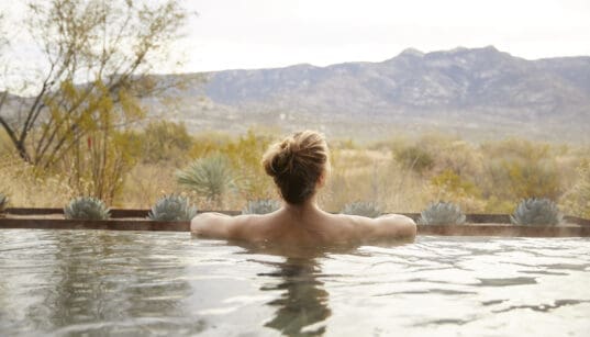 Guest relaxing in an outdoor soaking tub at Miraval Arizona Resort & Spa with desert plants and mountain views in the background.