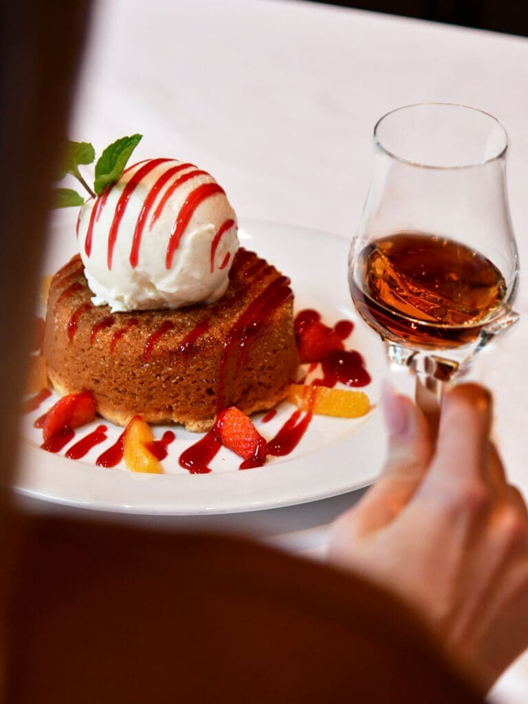 A person is holding a wine glass next to a plated Butter Cake served at Mastro’s steakhouse in Arizona.