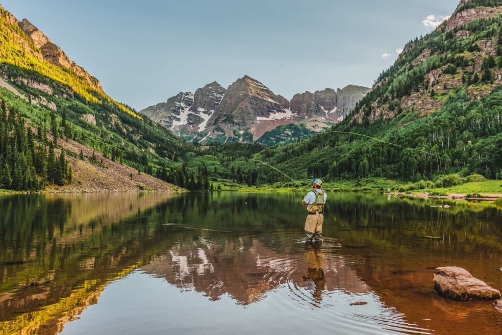 Fly fisherman standing in a mountain lake near Aspen, Colorado with views of the Maroon Bells and surrounding alpine scenery.