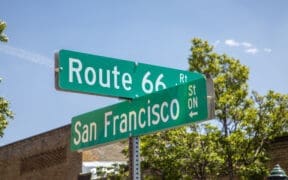 Route 66 and San Francisco Street signs in downtown Flagstaff, beneath a bright blue sky.