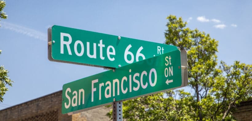 Route 66 and San Francisco Street signs in downtown Flagstaff, beneath a bright blue sky.