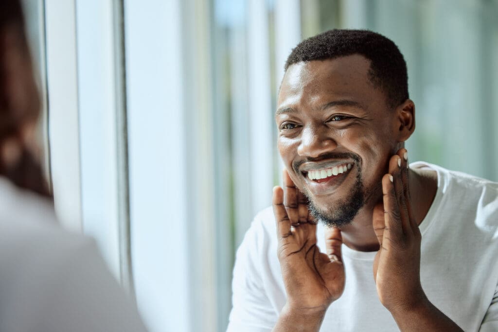 A smiling man looks at his reflection in a mirror while checking his skin as part of a daily skincare routine.