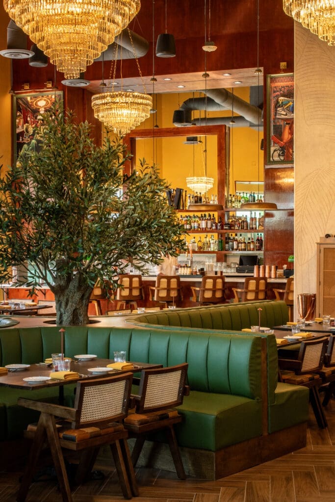 Dining room at NADU Phoenix featuring emerald green banquettes, crystal chandeliers, an olive tree centerpiece and a modern bar.