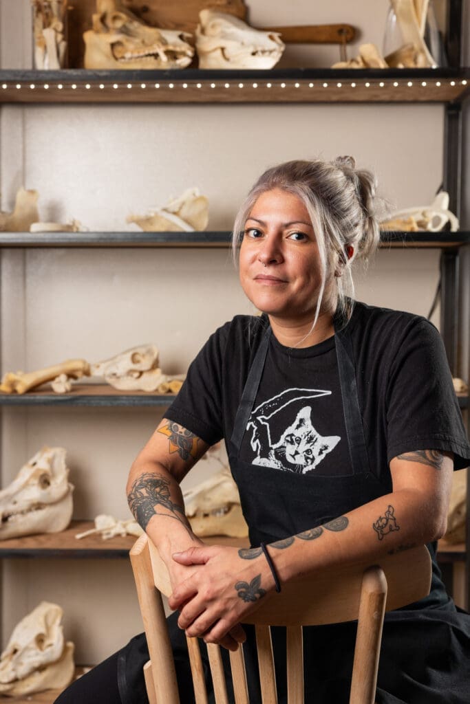 Chef Misti Norris sits in a chair inside a culinary workspace decorated with animal skull displays behind her.