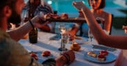Group of friends enjoying a fancy dinner with appetizers and sparkling wine by a pool at dusk during summer.