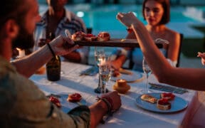 Group of friends enjoying a fancy dinner with appetizers and sparkling wine by a pool at dusk during summer.