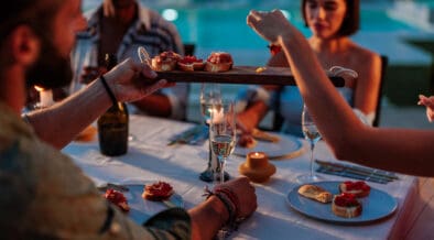Group of friends enjoying a fancy dinner with appetizers and sparkling wine by a pool at dusk during summer.