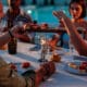 Group of friends enjoying a fancy dinner with appetizers and sparkling wine by a pool at dusk during summer.