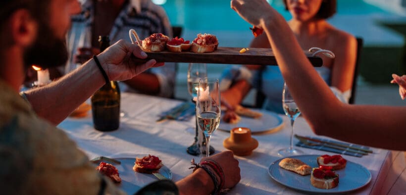 Group of friends enjoying a fancy dinner with appetizers and sparkling wine by a pool at dusk during summer.