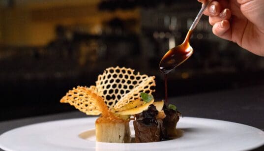 A plated dish at Atria in Flagstaff features wagyu, potato pavé and delicate garnishes as sauce is spooned over the dish.