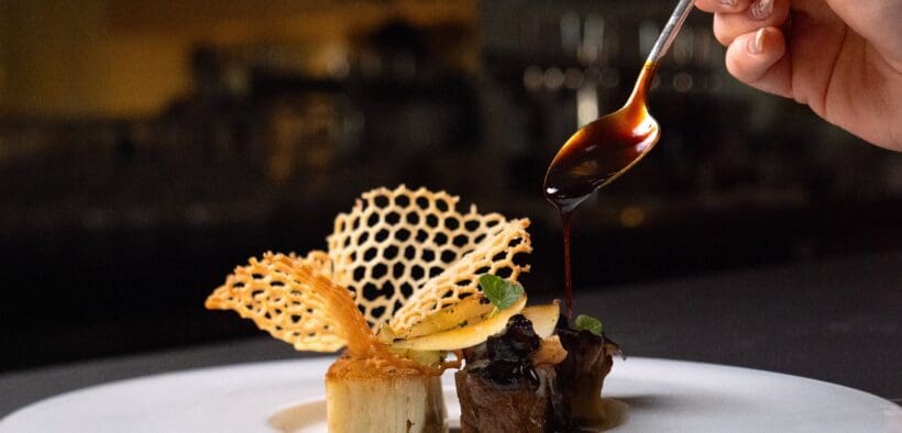 A plated dish at Atria in Flagstaff features wagyu, potato pavé and delicate garnishes as sauce is spooned over the dish.