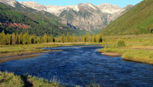River flowing through a green mountain valley beneath snow-dusted peaks near Telluride, Colorado.
