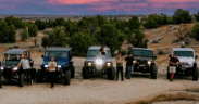 Off-road vehicles and drivers gather on rocky terrain beneath a colorful sunset sky in Farmington, New Mexico.