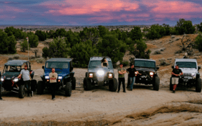 Off-road vehicles and drivers gather on rocky terrain beneath a colorful sunset sky in Farmington, New Mexico.