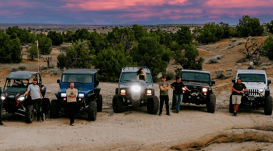 Off-road vehicles and drivers gather on rocky terrain beneath a colorful sunset sky in Farmington, New Mexico.