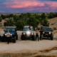 Off-road vehicles and drivers gather on rocky terrain beneath a colorful sunset sky in Farmington, New Mexico.