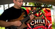 A man plays an electric guitar with the Las Cruces Hatch Chile Festival Logo sign in the background.