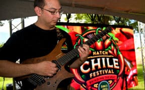A man plays an electric guitar with the Las Cruces Hatch Chile Festival Logo sign in the background.