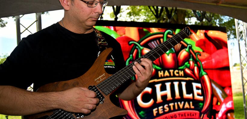 A man plays an electric guitar with the Las Cruces Hatch Chile Festival Logo sign in the background.