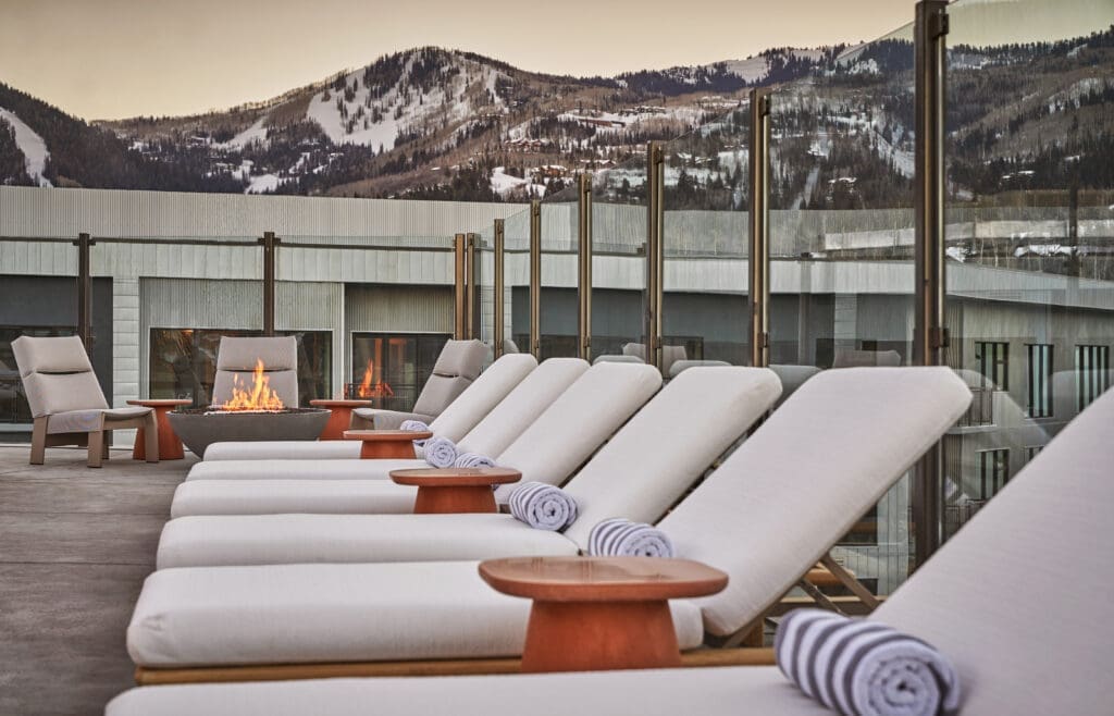 Lounge chairs and fire pits on an outdoor terrace at Pendry Park City with snow-covered mountains in the background.