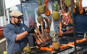 Chef preparing a large rack of meat alongside grilled cuts and pineapples at a previous RARE Steak Championship.