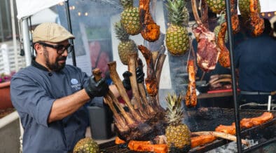 Chef preparing a large rack of meat alongside grilled cuts and pineapples at a previous RARE Steak Championship.